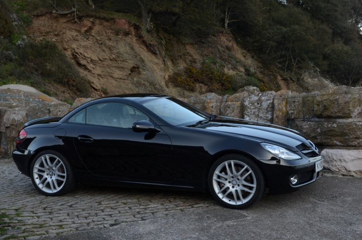 Show us your Mercedes! - Page 9 - Mercedes - PistonHeads - The image showcases a black convertible car parked on a cobblestone street. The vehicle is sleek, with its top down, revealing a spacious interior. The stonework of the street and the side of the car contrast beautifully, adding to the elegance of the scene. The rearview of the car and the mountains in the background create a picturesque setting. The car appears to be empty, parked and ready for a drive.