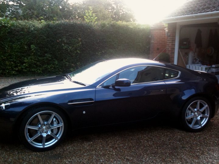 newby needs advise - Page 1 - Aston Martin - PistonHeads - The image depicts a sleek, black Aston Martin sports car parked in a driveway. The car is positioned at a slight angle facing to the left of the frame. It features a distinctive grille and Aston Martin emblem, indicative of luxury and high performance. The sunlight casts playful shadows across the car's glossy exterior. In the background, there's a glimpse of a red brick building, suggesting a residential or commercial area. The exterior of the driveway shows some greenery and concrete, adding to the ambience of a typical suburban setting.