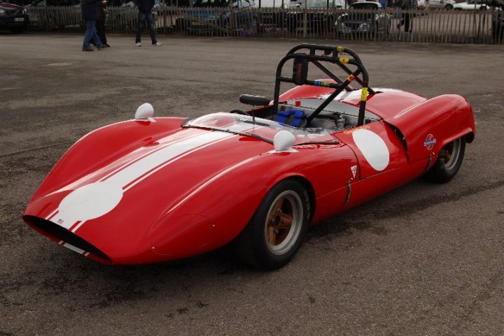 Pistonheads - The image shows a red sports car with a notable large white spot on its side, which could be seen as a star. The vehicle exhibits a classic design with a removable hardtop and side windows. The car is parked on a paved surface, and in the background, there are people walking, suggesting that this may be a public or populated area. The sports car has a sleek, contoured body, indicative of its high-performance capabilities.