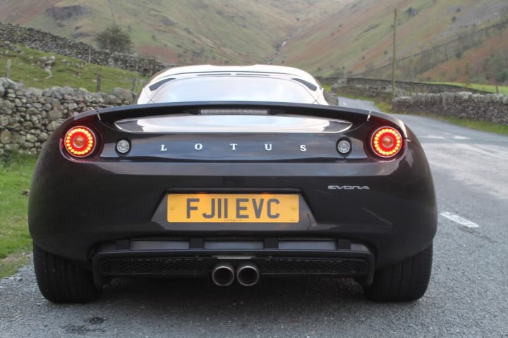 black evora spotted - Page 1 - Evora - PistonHeads - The image showcases a sleek and modern Lotus sports car on a winding street. The car's number plate reads "F JL EVC", indicating it might be on a track or a highway. The car is positioned on the side of the road, allowing for a clear view of its design and features. The landscape around the location is hilly and lush, adding a serene backdrop to the scene. This could possibly be during a sunset, given the warm lighting and the shadows cast by the car.