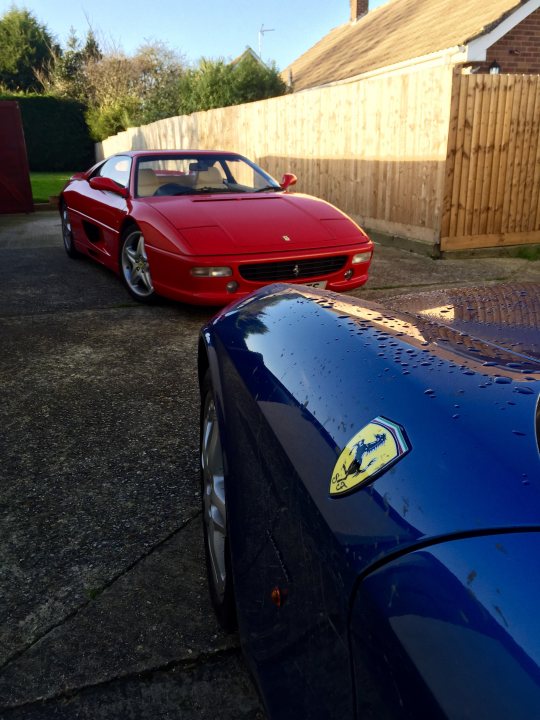 Christmas Day Drive - Page 1 - Ferrari V8 - PistonHeads - In the image, there are two luxury cars parked on a driveway next to a wooden fence. The first car is a striking red Ferrari with unique curved bodywork, while the second car is a sleek blue, its color accentuated by water droplets on its surface. Both cars are parked under the open sky, which suggests the photo was taken on a clear day. The positioning of the cars indicates they are parked parallel to each other along the curved fence. The red Ferrari's shiny exterior contrasts with the matte finish of the blue car. The wooden fence adds a rustic charm to the scene, suggesting a suburban or countryside setting. The phrase "It was like old times, just me, an F40, and the world" implies a sense of solitude and admiration for the Ferrari.