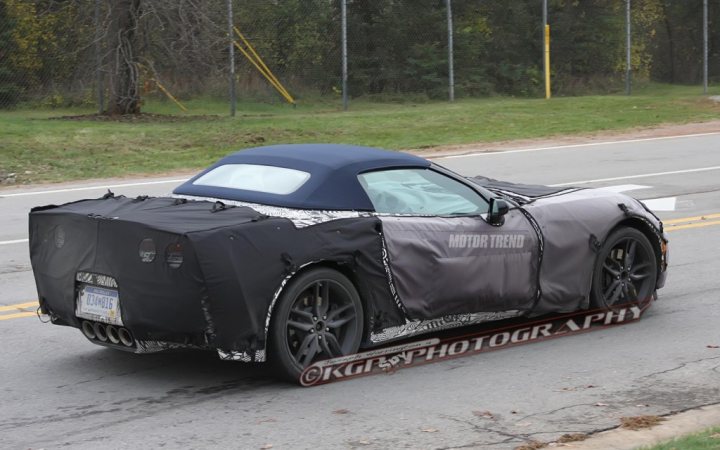 A car that is sitting in the grass - Pistonheads - The image shows a black, two-door convertible car partially covered with camouflage, suggesting it may be a prototype or a model under development. The car is in motion on a paved road, and while the windshield and rear window are visible, the body of the car is largely concealed due to the covering. The wheels and tires are exposed, showing standard road-ready design. A watermark at the bottom of the image indicates it is from "RoadBLOG Photography."