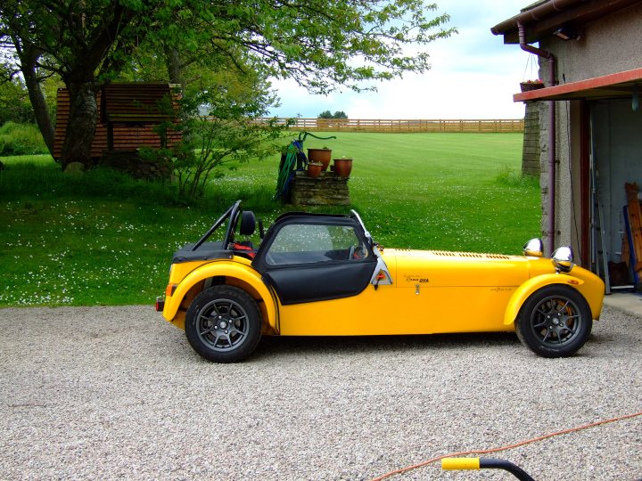 Not enough pictures on this forum - Page 34 - Caterham - PistonHeads - This image features a vibrant yellow and black two-seater convertible parked on a gravel driveway. The car has black accents and a sleek convertible top that is currently down. The vehicle is positioned in front of and to the left of a garage, and there are several objects around, including potted plants, an archway, and what appears to be gardening or storage equipment. The overall setting suggests a rural or suburban environment.