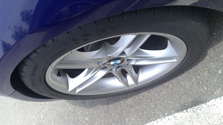 A close up of a car parked on a street - This image shows a close-up view of a car's wheel and tire. The tire appears to be a standard vehicle tire, approximately 245/450 size given the aspect ratio and width of the tread pattern. The wheel itself is multi-spoke, likely an aftermarket design rather than an original equipment manufacturer (OEM) part, with a silver or chrome finish. The tire has a visible tread pattern featuring multiple sipes, which are suggested by the indentations along the edges of the tread grooves. The overall condition of the wheel and tire suggests they are well-maintained, with no visible wear or damage.