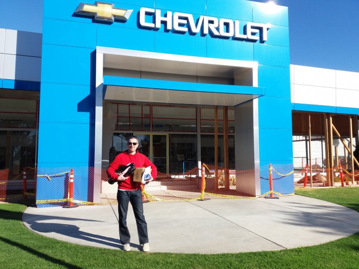 The £7700 Corvette C6 - Page 1 - Readers' Cars - PistonHeads - In the image, a man stands confidently in front of a large Chevrolet dealership. The man is dressed smartly in a red jacket and glasses, holding a box and a book, suggesting he might be presenting or showing the dealership something. The dealership itself is spacious and impressive, with a prominent blue and white sign proudly displaying the brand 'Chevrolet'. There's construction visible in the background, indicating that the dealership might be in the process of expanding or renovating. The cheerful surroundings and the man's relaxed stance suggest a friendly, welcoming atmosphere.