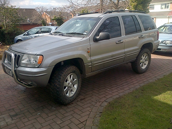 Grand Cherokee, any good? - Page 1 - Off Road - PistonHeads - The image shows a side view of a light-colored SUV parked on a driveway. The SUV has tinted windows, and the vehicle's wheels are visible. The driveway is equipped with brick paving. In the background, there are other parked cars, a house, and a tree, which suggests a residential area. There is no text visible in the image. The SUV appears to be well-maintained and is the main focus of the image.