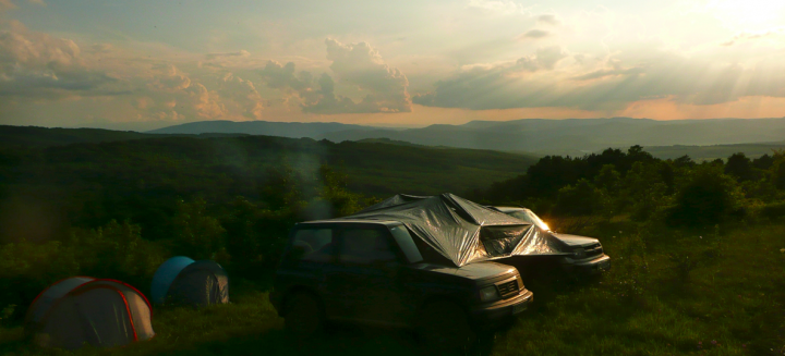 Little overnight trip to the Balkan range - Page 1 - Off Road - PistonHeads - The image captures a serene outdoor scene with two adventurous vehicles - a blue tent is set up on the left, perhaps serving as a temporary home, while on the right, a silver tent has been pitched, possibly providing a space for relaxation or an anticipation of an adventure ahead. The vehicles are parked on a grassy area, which stretches out to a lush, hillside terrain that rolls gently into the distance. The expansive sky overhead is painted with hues of orange and yellow, suggesting the time may be around dawn or dusk. The overall atmosphere is peaceful and tranquil, indicative of a brief pause during an adventurous journey.