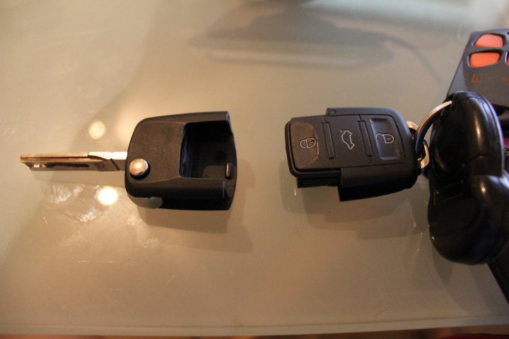 Charging remote keys ...remotely? - Page 1 - Rolls Royce & Bentley - PistonHeads - The image shows a pair of keys with a remote on a reflective surface. The keys are black and appear to be opening some devices. There is a remote control next to the keys, with three orange buttons visible on it. Both the keys and remote are laying on a table, and there is light reflected on the surface, likely from the camera flash or another source of light.