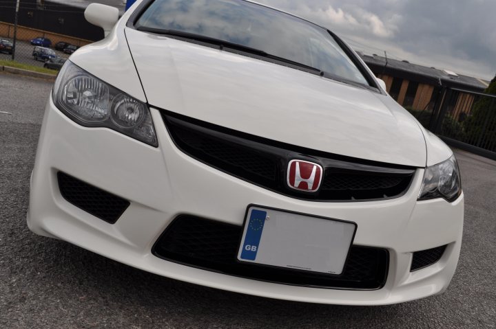 Import Pistonheads Type - The image shows the front view of a white Honda car parked. It has a distinctive front grille with the Honda logo in the center. There's a license plate fixed to a plate holder at the front. The car appears to be in good condition, and the parking lot provides a neutral background.