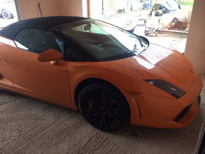 More pics of my LP560 - Page 1 - Gallardo/Huracan - PistonHeads - The image shows a bright orange convertible sports car parked indoors. The car's top is down, revealing a sleek interior structure that contrasts with the vehicle's vibrant exterior color. The wheels are black, complementing the car's body color. In the background, the scene outside of the structure features an assortment of other vehicles, which seem to be stored or parked.