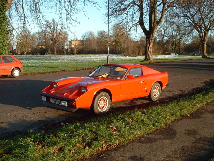 A red car is parked on the side of the road - Pistonheads - The image features a vibrant red classic Ferrari parked on the northern side of a street in a park-like setting. It's a bright day with clear skies, and there is a mix of leafless and lush trees, suggesting it might be fall. Another car, an orange Volkswagen Beetle-like vehicle, is parked in the background, adding a contrasting second subject. The surrounding leaves, indicative of autumn, add a charming seasonal touch to the scene.