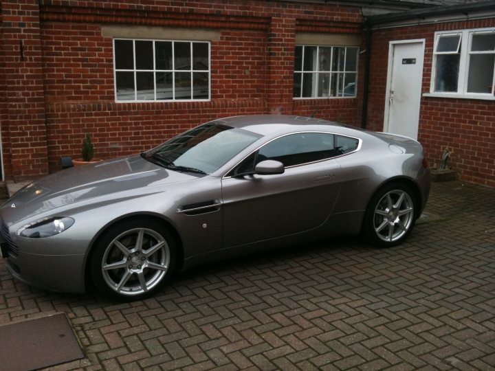 Vantage ownership? - Page 1 - Aston Martin - PistonHeads - The image features a striking gray Aston Martin sports car parked in front of a brick building. The car is positioned in front of the building, with its sleek design and distinctive grille facing towards the viewer. The brick building has a white door on its right and a window on the left, providing a contrasting backdrop for the car. The car's position slightly to the left of the frame adds a dynamic quality to the image, creating a sense of anticipation for what lies ahead. The clear visibility of the surroundings and the car itself suggests a bright, possibly overcast day.