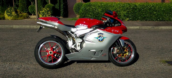 Pistonheads - The image shows a motorcycle parked on a paved street. The motorcycle is predominantly silver in color, with parts of it also featuring bright red and accents of black. The motorcycle is oriented towards the viewer, presenting its right side. The motorcycle model is not fully visible, but it is identified with a badge on the side. The background of the image features a clear sky, and there are green hedges and trees along the sidewalk, indicating a residential or possibly a semi-rural setting.