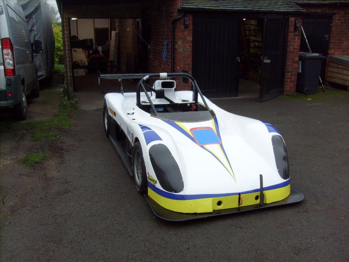 Casper goes to Romania - Page 1 - Radical - PistonHeads - The image shows a single-seat race car parked on an asphalt surface. The car is predominantly white and blue with a large yellow shape running down the center, reminiscent of a bird's beak. The vehicle features an open cockpit, and a roll bar is visible inside. It is parked in front of a brick building, indicating an indoor or semi-outdoor storage setting. A few handbags and personal items can be seen on the ground, and another car is visible in the background.