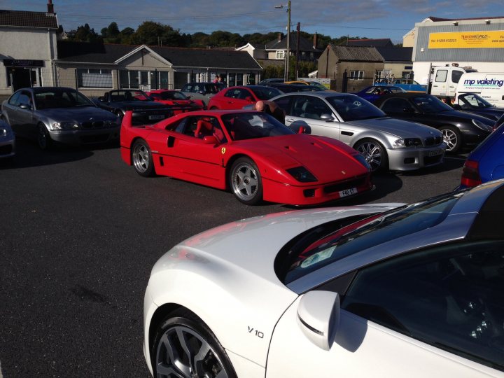 South West Wales Breakfast Meet - Page 143 - South Wales - PistonHeads - The image depicts a parking lot scene. An unusual red Ferrari, reminiscent of the F40, is parked in front of a white car. The Ferrari's bright color stands out from the otherwise somber setting. The white car's model is identifiable, showing that the image might be related to motoring events. Multiple other cars and a truck are scattered throughout the parking lot, creating a diverse backdrop for the main subjects. The setting suggests a public parking area, possibly near a building with a yellow and white sign that reads "Dell".