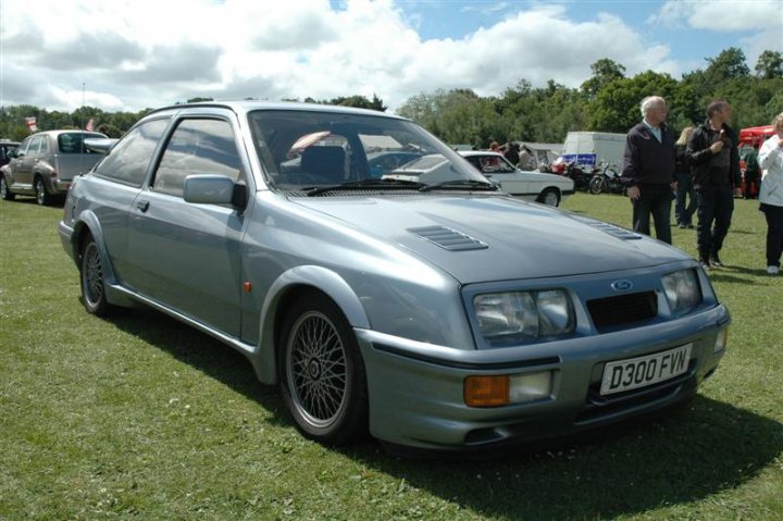 Fins and Chrome - Page 1 - North East - PistonHeads - The image depicts a silver, two-door hatchback car parked on a grassy field, possibly on show at a car exhibition. The car features a distinctive front bumper and a set of dark alloy wheels. In the background, there are spectators and other vehicles, indicating the car show setting. The sky overhead is overcast, suggesting an outdoor event taking place under cloudy conditions.