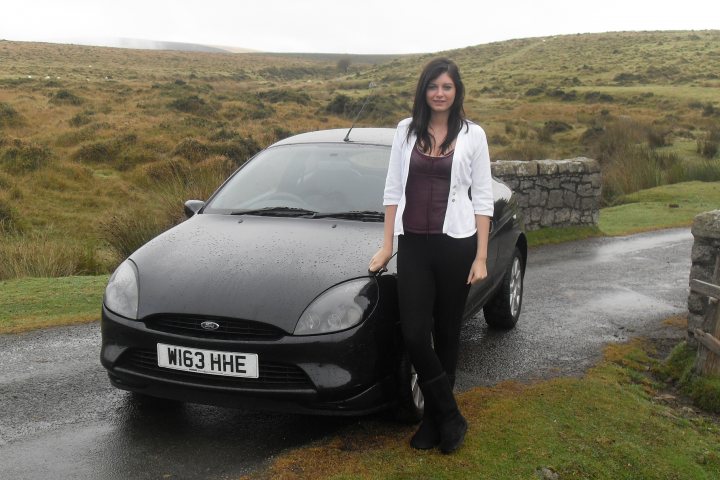 Puma - my first P&J - Page 1 - Readers' Cars - PistonHeads - In the image, a woman is standing next to a black car on a road. She is positioned behind the open door of the car, posing cheerfully for the photograph. The car has a license plate that reads "W53 HE." The backdrop features a pleasant scene with a grassy area and a hill. It appears to be a chilly day, as evidenced by the woman's smile and the romantic setting.