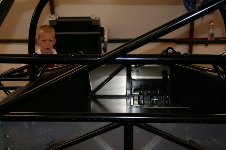 Ultima Carbon Cockpit Pistonheads - This image captures a moment inside a car shop. The main focus is a boy standing behind a black car frame. He seems to be inspecting or admiring the sleek frame of the car. The car frame is intricate, with a gray tube anchoring the center, suggesting a work in progress. The background of the image reveals a shop setting with tools and equipment, further reinforcing the car shop context. The boy is the only person in the frame, thus becoming the focal point of the image. The image evokes a sense of anticipation and curiosity, as the viewer wonders about the progress of the car's construction.