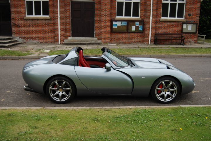 Random TVR Pics (Vol 2) - Page 44 - General TVR Stuff & Gossip - PistonHeads - The image is a photograph of an open-top sports car parked on the side of a street. The car is silver with red detailing, and it appears to be a two-door convertible with its top down. It has distinctive wheels and a sleek design that suggests speed and performance. In the background, there are buildings including a church, and the setting looks like an urban or suburban environment. There are no visible texts or logos in the image that provide additional context or branding.