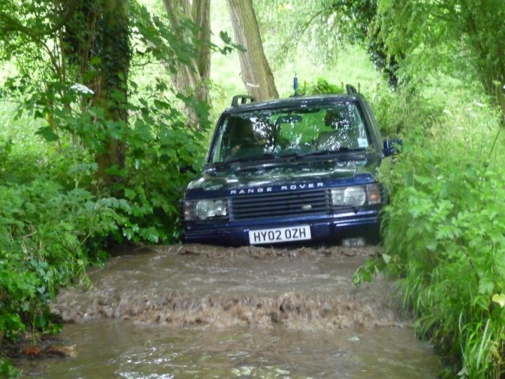 Show us your fording pics.... - Page 1 - Off Road - PistonHeads - The image depicts a dark blue Range Rover partially submerged in a river or flooded area, surrounded by trees and bushes. The vehicle's lights are on, suggesting it has been maneuvered there during daylight. The speed bump in the water is a notable feature, indicating that the flood water has risen to or above the barrier. The vehicle appears to be stuck, with its front end partially submerged, indicating it may have struggled during the flooding event.