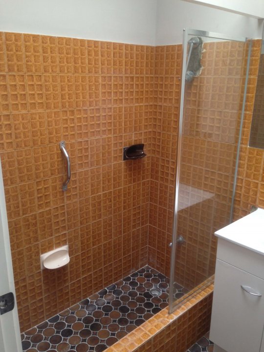 A bathroom with a toilet and a shower - Pistonheads - The image captures a compact, efficiently designed bathroom. The shower is enclosed with a clear glass door, and inside, there's a round dinghy hanging from the wall, ready for use. Adjacent to the shower, a small white sink is mounted on the wall. The floor of the bathroom is covered in orange and brown tiles, adding a warm and inviting touch to the space. A small window punctuates one of the walls, providing a hint of natural light.