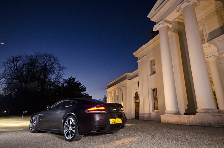 My V12V photographed by a talented photographer (not me) - Page 1 - Aston Martin - PistonHeads - This is a photograph showcasing the back of a black car, presumably a modern sports car given the taillight design and the sleek silhouette. The car is parked in front of an architectural structure, likely a building or a viewing tower, with large circular pillars that are detailed with ornate carvings, visible in the background. The overall setting is an outdoor night scene, with the car's tail and license plate illuminated, a soft gradient of the blue sky to night extending over the building, and the landscape silhouetted against the darkness. There are no visible people in the image.