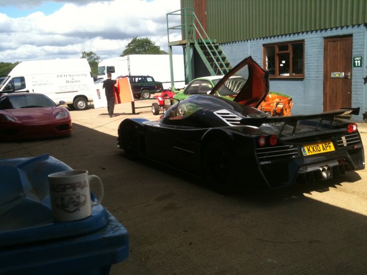 Pistonheads Ultima Competitors - This image captures a bustling scene in a parking lot next to a building with a green roof. Black sports cars, vibrant with red and green accents, dominate the scene. One striking car with its hood open reveals a sleek engine, while another car showcases its yellow license plate and a red sunroof. Amidst the dynamic setting, coffee cups are visible on a side table, suggesting frequent work or meetings in this location.