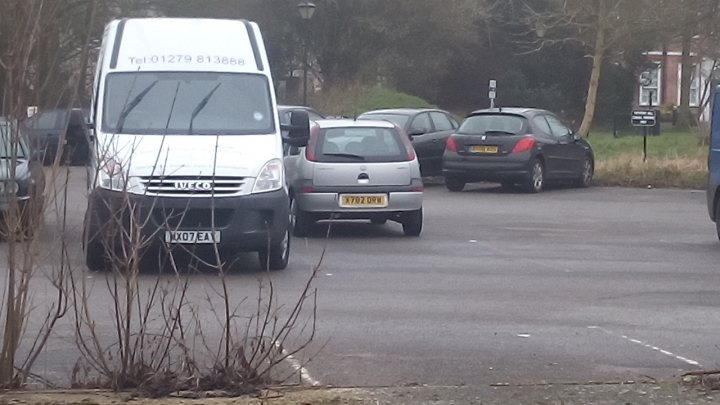 Lets play the Photo Location Game! Vol 3 - Page 48 - General Gassing - PistonHeads - In the center of the image, a blue van is parked diagonally in a parking lot, with its front facing the top right corner of the picture. Around the van, several other vehicles can be seen, including a white truck on the other side of the blue van, a black compact car, and a silver sedan. Behind these vehicles, grass and a building hint that the parking lot is located in a suburban or rural area. The sky is visible in the top left corner of the image, suggesting that the photo was taken during the day.