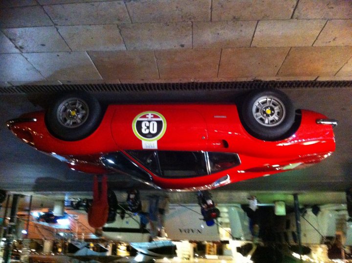 Driving France - Page 4 - Chimaera - PistonHeads - The image captures a scene from an underground parking garage. The main subject is a highly raised red sports car that is turned upside down for inspection or maintenance. The car is positioned under a large cement patch in the ceiling, which casts a strong shadow on the car. The photo is taken from a low angle, emphasizing the perspective of the car's odd disposition. This view makes the car appear like a Venn diagram, with the borders of its usual positions worryingly close to each other. The rest of the parking garage is largely empty, with only a few tables scattered in the background, adding a sense of depth and scale to the scene.