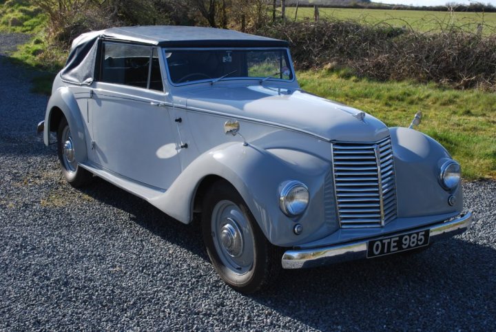 Anyone remember this car from the 1950s or 60s? - Page 1 - North West - PistonHeads - The image showcases a vintage car with a sleek design, dominating the scene. Its silver paint gleams under the light, reflecting the surrounding environment. The car appears to be stationary on a gravelled area, which provides a rustic blend with the green, leaf-filled field located in the background. The windshield of the car is adorned with tall grasses around the front, and a solitary lamppost stands sentinel to the right. The tranquil setting exudes a serene charm, contrasting the vintage car's classic elegance.