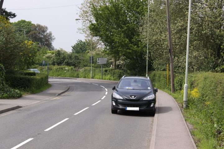 Parking Issue - Page 1 - Speed, Plod & the Law - PistonHeads - This image depicts a gray car driving on a paved road. The car is on the side of the road, seemingly making a right turn onto a side street. There are trees along both sides of the road, providing a canopy of green foliage. The sky appears to be clear and blue, suggesting fair weather. There are no visible buildings or distinctive landmarks, indicating that the car is in a more suburban or residential area. The overall scene is quiet and peaceful, with no other vehicles visible in the immediate vicinity.