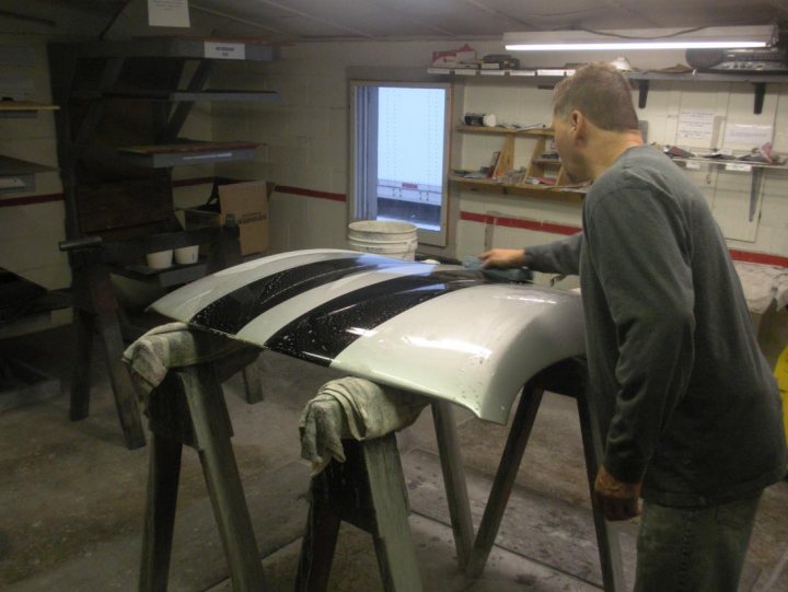 New top - Page 1 - Vipers - PistonHeads - This image captures a moment in a workshop where a man is painting a distinctive black and white striped design on a white surfboard. The man, standing on the right side of the image, is using a paint sprayer, meticulously applying the stripes to the surfboard. The surfboard is on a wooden workbench, which is equipped with a dustpan. In the background on the left, two wooden chairs can be seen, possibly for the comfort of the artist. The window on the wall in the background suggests that the workshop is well-lit, indicating the man's commitment to precision in his art.