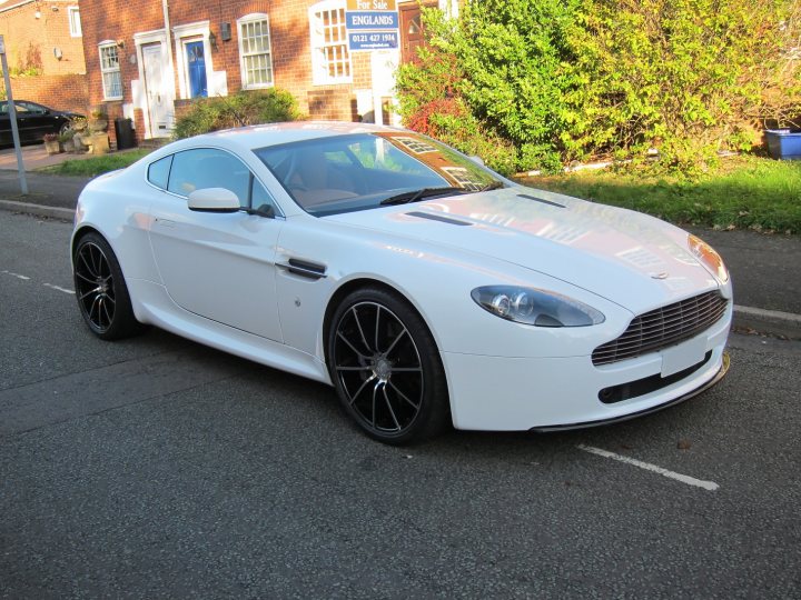 Show me your wrapped v8v - Page 1 - Aston Martin - PistonHeads - The image depicts a sleek, white sports car parked on a city street. The car, which appears to be a Jaguar, features a striking design with a front license plate and a distinctive grille design. The vehicle is parked partially on the pavement and partially on a line painted on the street, indicating it is parallel parked. The setting is urban, with clear visibility of the facade of a brick building with two front doors across the street. The lighting suggests it is daytime, and there are trees in the background, providing a contrast to the cityscape.