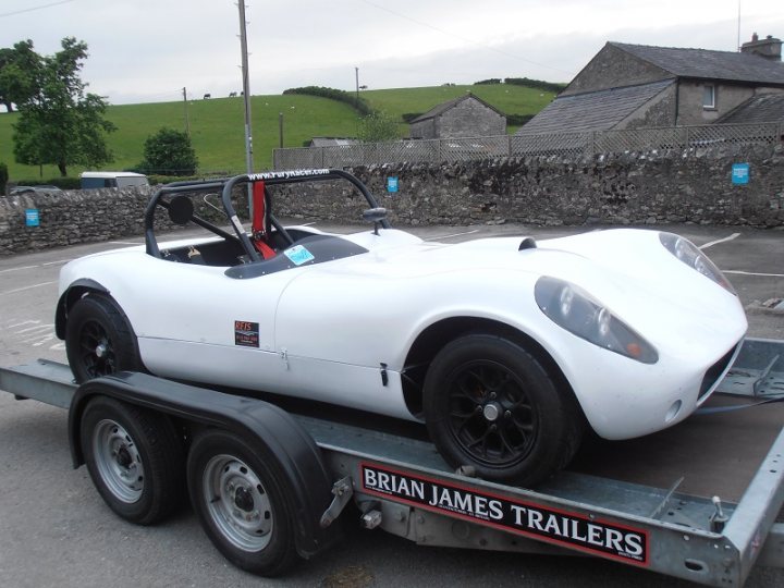 whats an ideal budget trailer for clubsport/prosport etc? - Page 1 - Radical - PistonHeads - The image features a white convertible car being towed by a trailer. The car is positioned facing forward on the trailer. The trailer, bearing the sign "Brian James Trailers," is parked in a lot next to a brick wall. In the background, there are trees and houses, suggesting a suburban setting.