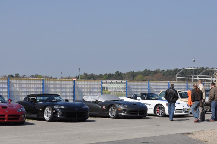 Pistonheads Jim Hockenheimring Clark - The image shows a row of cars parked in a lot adjacent to a tree line. There are four cars in total, including one that is divided into two visible sections. Each car is parked side by side, and there are people walking in the vicinity, possibly inspecting the vehicles. The setting seems to be a clear day, and the cars appear to be luxury or performance types, judging by their design and size. The overall atmosphere is relaxed, with no immediate evidence of a formal event.