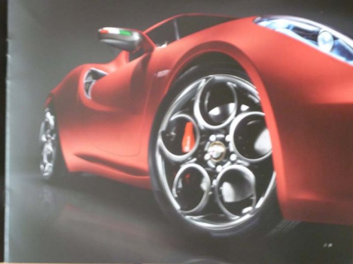 Received an Alfa 4C brochure through the post today. - Page 1 - General Gassing - PistonHeads - The image features a red sports car depicted from an angle that showcases the vehicle's side profile and the front wheel. The focus is clearly on the car's design, particularly the shiny metal spokes of the wheel and the details of its metallic surfaces. The car appears to be a high-performance model, with a streamlined silhouette and round, multispoke wheels. The color composition is monochromatic with the red of the car providing a stark contrast against the black road surface and the dark background. The overall style of the image is glossy and polished, emphasizing the car's sleek lines and reflective surfaces.