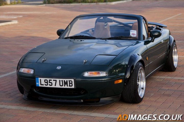 Buying an mx-5: What do i need to know? - Page 3 - Mazda MX5/Eunos/Miata - PistonHeads - The image captures a vibrant blue sports car parked in a lot. The car, labeled with the registration "L957 UMB," is sleek with a convertible top down, allowing for an internal view. The windshield is clean and clear, reflecting the open sky above. The surrounding area includes a paved lot and a hint of a building in the background, giving a sense of location.