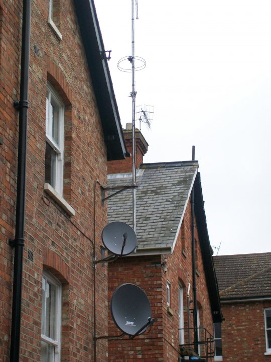 Pistonheads - The image captures a brick building with a distinctive array of satellite dishes affixed to its exterior. It appears to be a residential structure, possibly an apartment building, given its multi-storied facade. The building's brickwork displays a mix of red and brown hues, providing a warm texture to the scene. The sky in the background is overcast, casting a soft light that contrasts with the darker brickwork. The image does not contain any visible texts or distinctive features that would provide additional context about the location or the building itself.