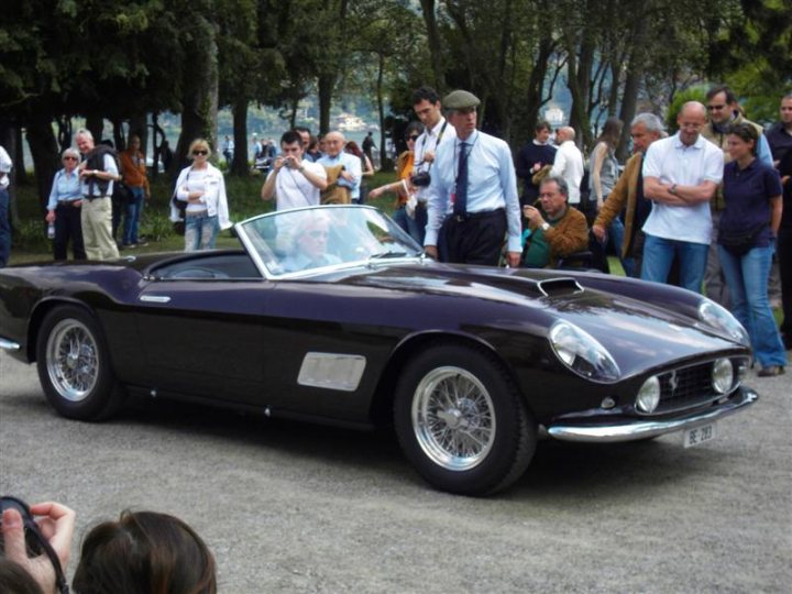 Villa Concoursporn Pistonheads Deste - The image depicts an elegant black sports car parked on a gravel area, where a crowd of people is gathered. The sports car is an early model monoposto and one of its most striking features is a glass windshield which wraps down to a low level at the back. The vehicle is distinguished by its silver hubcaps that add a touch of sophistication to its overall look. The scene is set outdoors, perhaps near a body of water or a forested area, indicated by the backdrop. In celebration of the image, spectators and photographers alike are seen admiring the antique vehicle.