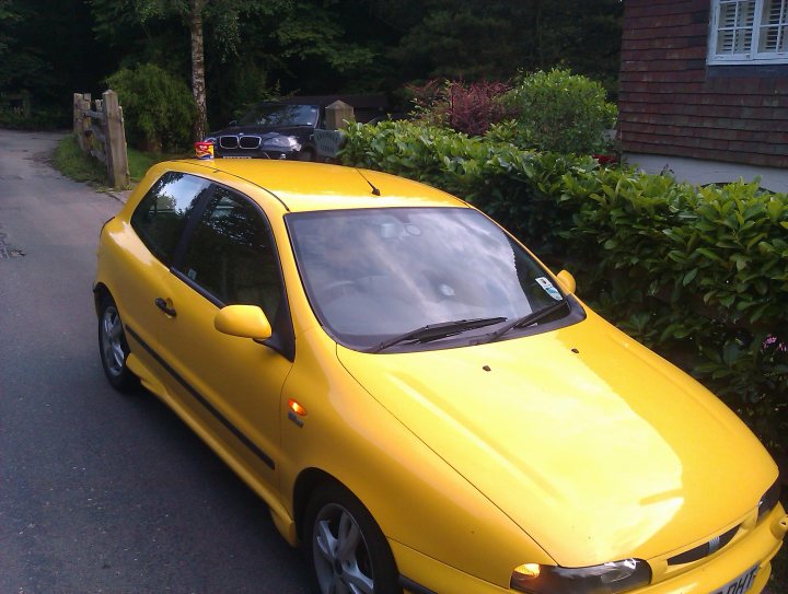 Lets play the Photo Location Game! - Page 12 - General Gassing - PistonHeads - The image features a vibrant yellow four-door hatchback car parked by the side of a road. The car is parked under a large tree with lush green leaves, providing shade. In the background, there is a house with a red brick facade partially visible. The road beside the car is relatively quiet with minimal traffic, suggesting it might be a residential area. The car itself appears to be in good condition, indicating it might be a common mode of transportation in this area.