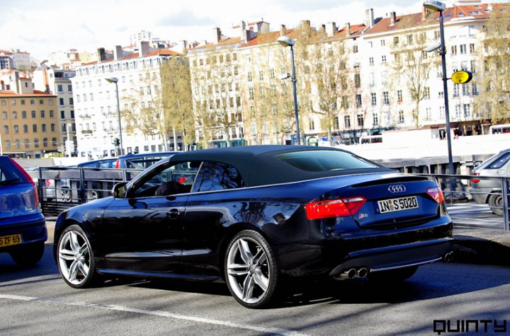 Debut Set October Droptop Audi Pistonheads - The image portrays a black sports coupe parked on the side of a road. The car is positioned in front of a building that appears to be a multi-story residential complex. There are two other cars in the adjacent lane. To the right, a traffic light can be seen, contributing to an urban setting. The sky is visible in the background, and the overall lighting suggests an overcast day.