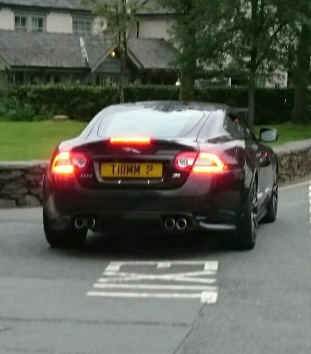 What C124PPY personalised plates have you seen recently? - Page 497 - General Gassing - PistonHeads - The image is a photograph of a black sports car parked in front of a building with a stone facade. The vehicle has a sleek design, featuring a large front grille and angular headlights. It's marked with the license plate "K12 MW" and bears a registration plate that reads "TIMOTHY 2." The car is equipped with dual exhaust pipes at the rear. The setting suggests it might be in a residential area or near a business establishment due to the presence of a parking spot designated by a white line. The photo appears to have been taken during the day under natural lighting conditions.