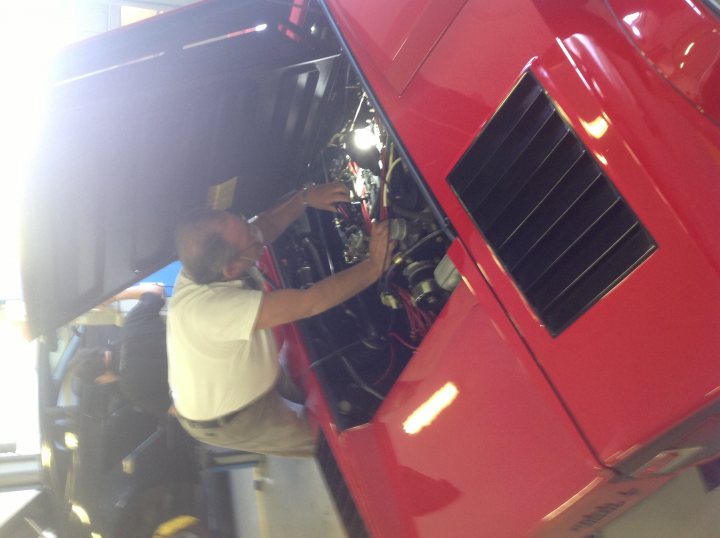 Scud Prices UP  - Page 5 - Ferrari V8 - PistonHeads - This image depicts a person bent over in a garage or workshop setting, working on the engine of a red car. The person appears to be male, wearing a white shirt and brown pants, and is busily adjusting or inspecting some component of the vehicle's under carriage. The car's hood is raised, revealing the engine inside, and the interior of the engine bay is visible in the background. The focus of the image is on the interaction between the person and the vehicle, capturing a moment of technical engagement in the auto repair context.