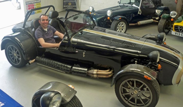 Driving Pistonheads Set Pls Broad - In the image, a man is sitting in a three-wheeled vehicle inside a garage or showroom. The vehicle has a sleek design with a black and silver body and large silver wheels. It appears to be a vintage or custom car, possibly a car that's been modified or has a unique design. The man is smiling, suggesting he is enjoying the ride. In the background, there are other smaller vehicles, indicating that this might be a place where custom or unusual cars are showcased or worked on, although their specific makes or models aren't discernible.