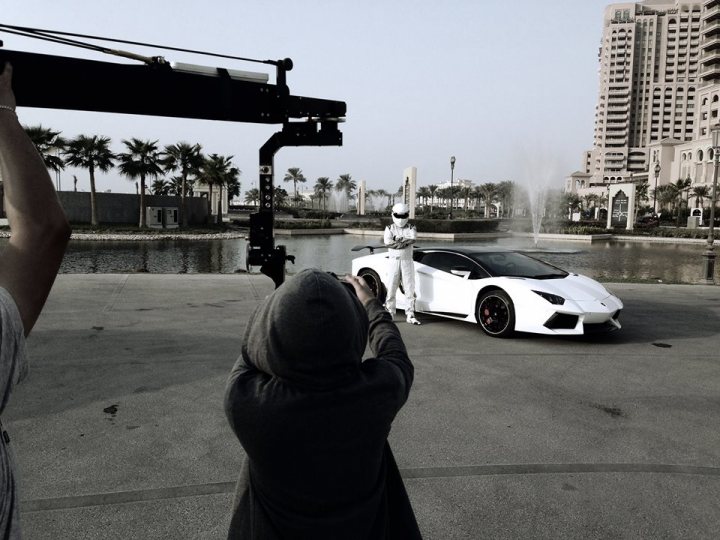 A man standing on the side of a road next to a car - Pistonheads - In this image, a white sports car is parked in a lot, with tall buildings and palm trees in the background. A person is seen taking a photograph of the car from a high angle, standing by a crane. In the foreground, another individual appears to be posing or interacting with the photographer, wearing a white jumpsuit. The atmosphere suggests an outdoor photography session during the day.