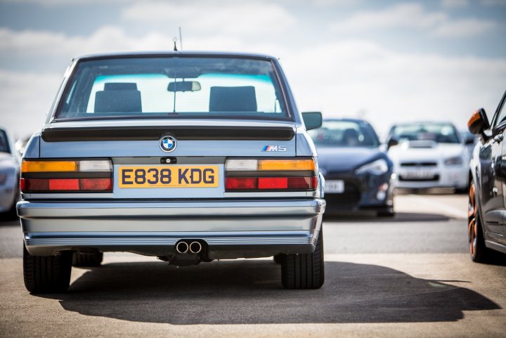 1987 BMW (E28) M5 - Page 1 - Readers' Cars - PistonHeads - The image captures a scene on a road where several cars are parked. In focus, there's a distinctive blue and black BMW, which stands out due to its unique color combination. The license plate of the BMW is clearly visible and reads "E83 KDG." The car's design elements, including the distinctive BMW logo on the hood, are discernable. In the background, the silhouettes of other cars can be seen, adding depth to the image. The sky in the background appears to be clear, suggesting an open-air environment.