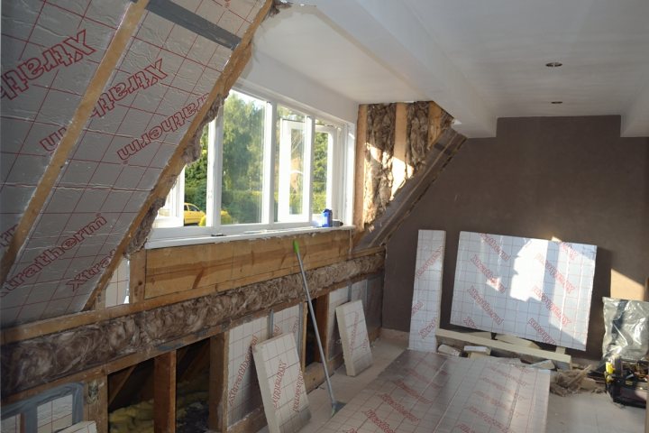 Insulating a dormer house - Page 2 - Homes, Gardens and DIY - PistonHeads - The image shows a room that is currently under renovation. The bare foundation and ceiling exposed the underneath structure of the room. The windows and frame around them indicate that the walls have been partially removed, and the bottom floor section also appears to be in the process of being worked on. A combination of construction supplies, tools, and three large, angled pieces of sheet metal suggests that some sort of structural reinforcement or skinc work is being performed. The additional presence of a ladder and power tool further indicates an ongoing construction project.