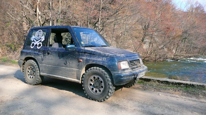 Winters over... Big chunky proper tyres back on... - Page 1 - Off Road - PistonHeads - The image shows a black SUV parked on the edge of a dirt road next to a river. The car appears to be old and battered, with visible mud splatters on the body. The windows are partially rolled down, suggesting that the car was recently driven on a muddy trail. Beyond the vehicle, there is a tranquil scene of a gentle river flowing through a forested area. It's a picturesque outdoor setting with a clear sky, which contrasts with the rugged appearance of the SUV.