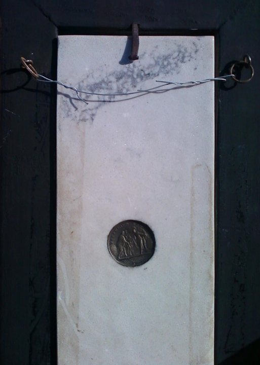 The image displays a commemorative plaque with a metal medallion affixed to it. The plaque itself seems to be made of a coarse white fabric or paper, mounted within a black frame that appears to be weathered. A stain, possibly of water or another substance, is visible on the plaque, creating small patches of dark discoloration. 

The metal medallion is egg-shaped, with the top curving smoothly over a lower, flatter section. The central part of the medallion features an embossed image of several figures, possibly a man, woman, and child, who seem to be interacting, but the exact content of the image is not clear. The background of this metallic embossment appears plain and unadorned. The medallion is held in place by ties, including what might be a clasp at the top and additional support bars running through it. 

The plaque is framed with a simple metal border and faces a dark background, which contrasts with the white of the plaque. The style of the image is a straightforward photograph, capturing this object in a way that focuses on the details of the plaque and medallion.