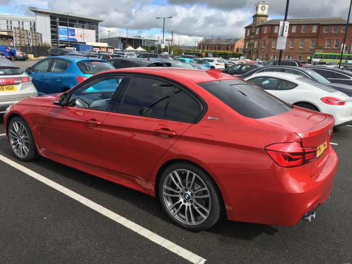 BMW 330e ordered... - Page 1 - EV and Alternative Fuels - PistonHeads - The image features a red BMW parked in a large parking lot, which is filled with various cars. The BMW car is sleek and modern, indicating it is likely of a newer model. In the background, a clock tower can be seen, suggesting the location might be a town center or a similar area with public buildings. The scene seems to capture a typical day in a bustling urban environment with the car appearing as if ready for a drive.